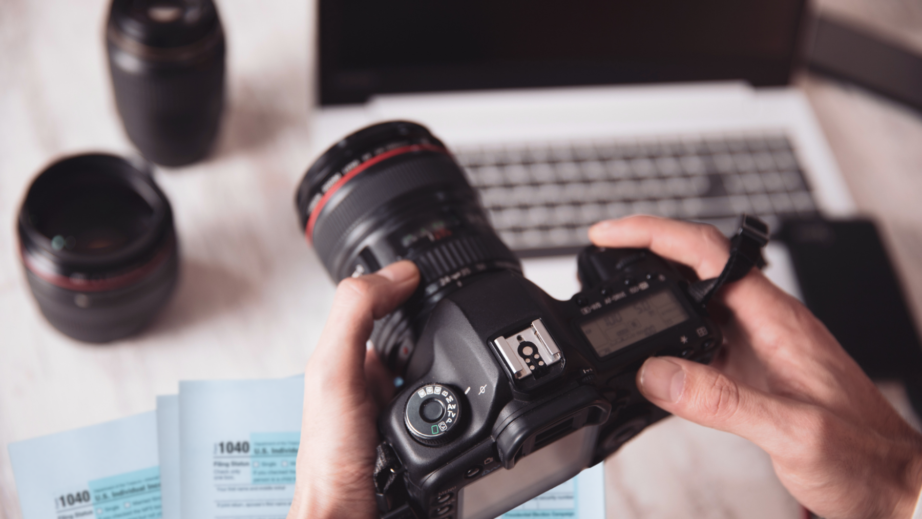 ax tips for freelancers illustrated by a photographer holding a camera above a laptop, 1040 forms, receipts, and a calculator on a desk.